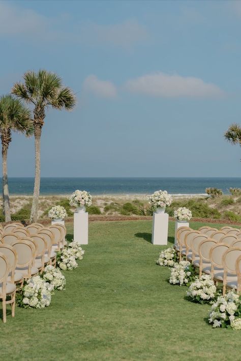 Outdoor wedding ceremony with white chairs facing the ocean. The aisle is decorated with white flowers and palm trees line the background. Beach Ceremony Florals, Beach Wedding Aisle Decor, Dock Wedding, Sea Island Wedding, Waterfront Wedding Ceremony, Wedding Columns, Wedding Bali, Wedding Ceremony Setup, Beach Wedding Aisles