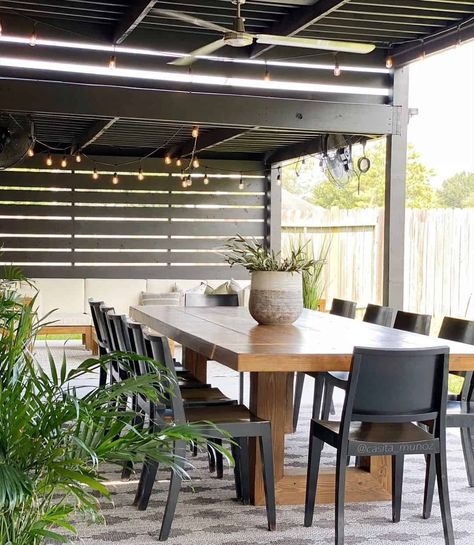 A black pergola is one of the hottest outdoor patio ideas and provides shade over the top of the wooden picnic table. Several black wooden chairs are against the table with a lovely flower pot in the middle. A white three-blade ceiling fan hangs off the pergola with decorative outdoor lights adding charm. A large gray and tan area rug sits in the middle underneath the table for comfort. Black Wooden Chairs, Enclosed Patio Ideas, Summer Patio Decor, Black Pergola, Pergola Outdoor, Small Outdoor Patios, Wooden Picnic Tables, Tan Area Rug, Wicker Coffee Table