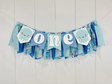 "HIGHCHAIR BANNER: ------------------------------ This handcrafted banner is the perfect piece for a Under The Sea 1st Birthday party! This eye-catching pennant flag banner is fit for many purposes. Perfect for a highchair, party decoration, cake smash photo shoot, or photo prop.  This listing includes both ONE BANNER AND FABRIC GARLAND.  Review sizing and description below. ONE BANNER: - Made from 65 lbs card stock paper. - Each paper pennant is 4.25\" in width across and 5.0\" in length down.  - TOTAL LENGTH OF PAPER BANNER is approximately 22 inches. FABRIC GARLAND: - Cotton fabrics tied into tassels and are hung behind the ONE banner.   - TOTAL LENGTH OF FABRIC GARLAND measures 2.5 ft across in width and 10 inches down in length. COLORS: - blues, teals NOTE: - Please measure to ensure Sea Creature Party, First Cake Smash, Under The Sea 1st Birthday, Paper Pennant, High Chair Decorations, 1st Birthday Boy, Birthday Highchair, Ocean Birthday, Cricut Baby