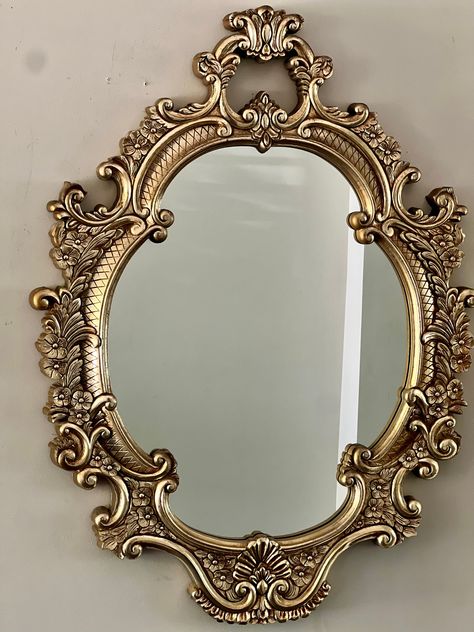 Iconic in its design and reminiscent of Old-Hollywood glamour, this ornately carved, oval, 1960s gold gilt mirror glams up your wall lending an inherent sense of luxury to any space.  Good vintage condition with gilt having acquired a warm patina. No cracks of any kind. Gorgeous Gold Gilt Heavy Plastic Composite Frame. Some visible shading and wear of gold gilt paint on frame and outer sides, which only adds glamour to the warm patina. One clamp for hanging the mirror is broken but the mirror ca Vintage Mirrors Small, Gold Princess Mirror, Circle Vintage Mirror, Ornate Wall Mirror, French Gold Mirror, Gold Accent Pieces, Vintage Gold Decor, Gold Apartment Aesthetic, Ornate Bathroom Mirror