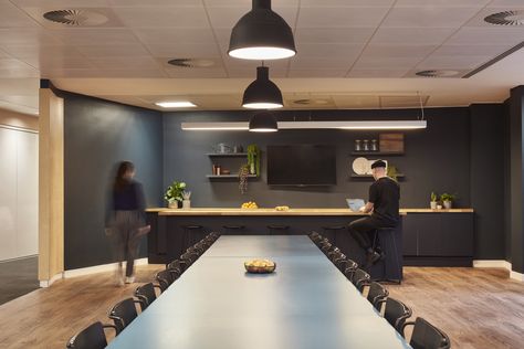 Two employees are at the dry bar in the Octagon Sports and Entertainment Network’s offices in London. One is on his computer sat at a high stool and one is walking to get herself a drink.

The walls and kitchen cabinets are dark blue and there are shelves styled with plants and crockery. There is a large screen for the team to gather, have refreshments and watch sports together and a large meeting table with chairs around it. Marketing Agency Office, Shared Workspace, Agency Office, Collaborative Workspace, Sports Marketing, Dry Bar, Social Space, Design Practice, Friday Afternoon