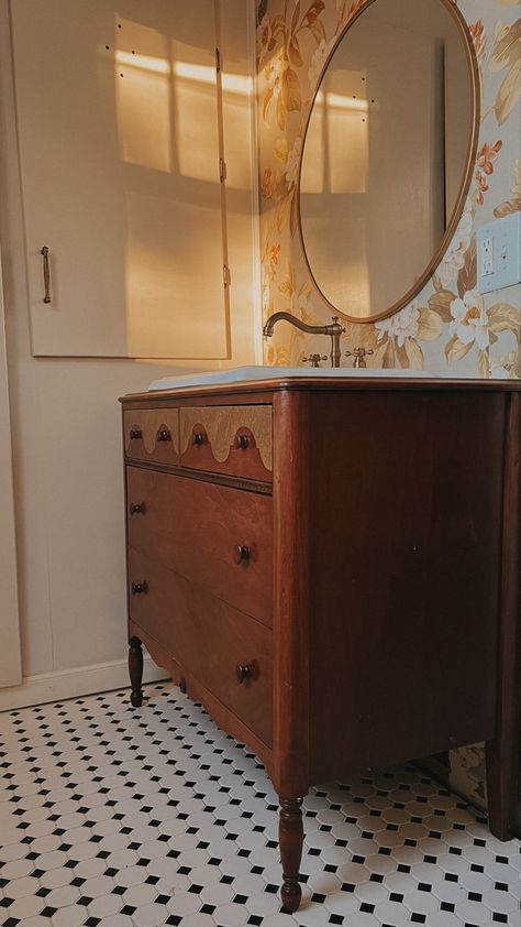 Wooden dresser, turned into a bathroom sink, vanity with floral wallpaper in the background, brass hardware, and a black and white checkered floor Vintage Vanity Bathroom Sink, Vintage Wooden Bathroom Vanity, Dresser To Sink Vanity, Bathroom Vanity From Old Dresser, Vintage Dresser Bathroom Vanity, Dresser As Vanity, Dresser In Bathroom, Antique Dresser Bathroom Vanity, Dresser To Bathroom Vanity