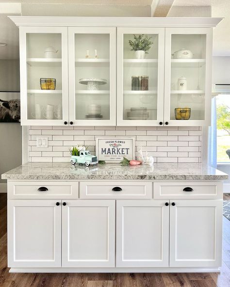 This neutral room features white kitchen cabinets with glass doors on the top section. A white subway tile backsplash contrasts with the gray marble countertop featuring rustic decor such as a wooden sign and spring themed ornaments. Glass Door Upper Kitchen Cabinets, Glass Front Kitchen Cabinets Ideas, Upper Kitchen Cabinets With Glass Doors, White Kitchen Cabinets With Glass Doors, Glass Kitchen Cabinets Decor, Kitchen Cabinets With Glass Doors, Glass Cabinet Decor, Glass Front Kitchen Cabinets, Renovate Kitchen