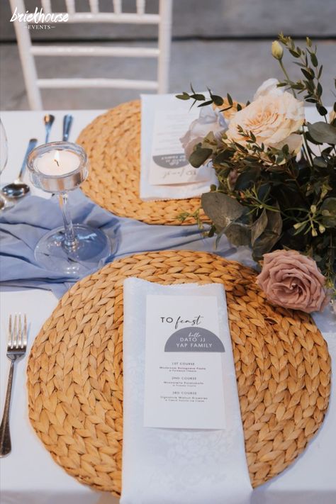 Wedding guest table arrangement with rattan placemats and blue runner decorated with flower centerpiece candles and glassware. Wedding Woven Placemats, Wicker Placemats Wedding, Rattan Placemats Table Settings, Wedding Table Set Up Ideas, Table Set Up Ideas, Dusty Blue Wedding Table, Centerpiece Candles, Blue Wedding Table, Wedding Themes Ideas