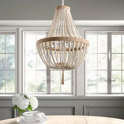 This pendant light showcases a breezy geometric design with a blend of metal and rope materials for a boho look in your space. It features two circular frames adorned with strands of wooden beads, creating an empire-like shade for an elegant yet rustic ambiance. A large circular bead dangles from the shade, adding a cohesive visual element. With four candelabra-style bulb sockets, this pendant light casts an ambient glow in your dining room, entryway, or living space. It's compatible with sloped Boho Chandelier Dining Room, Coastal Chandelier Dining Room, Beaded Chandelier Dining Room, Beachy Chandelier, White Beaded Chandelier, White Bead Chandelier, Lana Wood, Beaded Light Fixture, Coastal Light Fixtures