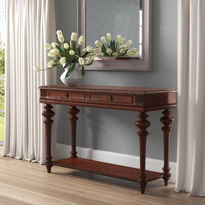 Defined by its turned trumpet legs and carved accents, this classic console table brings a touch of traditional character to your living room or entryway. Its rectangular top and lower open shelf provide the perfect place to display framed photos, potted plants, and a basket of magazines, while its two drawers offer concealed storage for odds and ends. Crafted from a blend of solid and manufactured wood, this versatile design sports a warm brown finish that pairs easily with most colour palettes Consoles In Living Room, Console For Entrance, Entrance Console Design, Classical Console, Heritage Table, Classical Room, Traditional Console Table, Shoot Moodboard, Classic Console Table