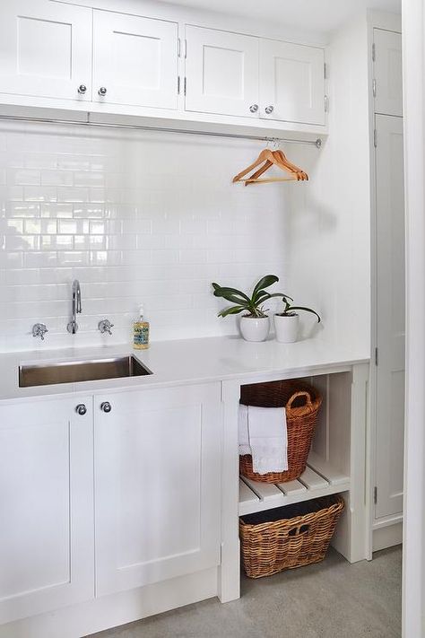 Monochromatic white laundry room boasts white slatted shelves holding woven baskets beside white cabinets adorning polished nickel knobs and a white quartz countertop framing a stainless steel sink positioned below a wall mounted polished nickel faucet kit fixed to a white subway tiled backsplash beneath a tension rod drying rack mounted under white overhead cabinets. Unfinished Basement Laundry, Utility Room Storage, Laundry Room Storage Shelves, White Laundry Rooms, Small Laundry Room Organization, Room Storage Diy, Basement Laundry Room, Laundry Room Flooring, Laundry Room Sink