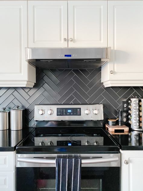 A kitchen with a black herringbone subway tile backsplash. The cabinets are white adding a gorgeous contrast. There is a black and stainless steel stove in the centre of the photo. There are also some natural wood accents and stainless steel canisters and spice rack on shiny black counters. Charcoal Herringbone Backsplash, Black And White Subway Tile Kitchen, Back Splash Ideas For Kitchen With Black Counter Top, Kitchen Backsplash Dark Grout, Black Backsplash With White Cabinets, White Kitchen Cabinets With Black Backsplash, White Kitchen Black Backsplash, Black Backsplash Kitchen White Cabinets, White Cabinets Black Backsplash