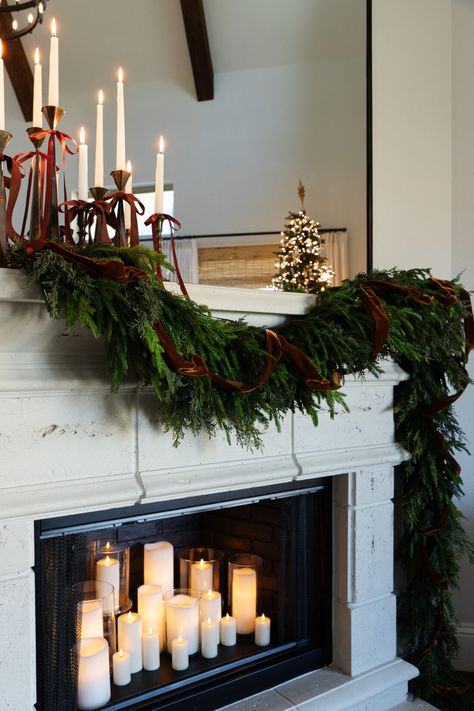 layered Christmas garland on fireplace mantle with brown velvet ribbon. Christmas bow trend using taper candles and brass candlesticks.  Lots of candles in the fireplace. Garland On Bookshelves, Christmas Garland On Fireplace, Candle Sticks On Mantle, Garland On Fireplace, Velvet Ribbon Christmas, 2024 Living Room, Garland Mantle, Bow Trend, Living Room Christmas Decor