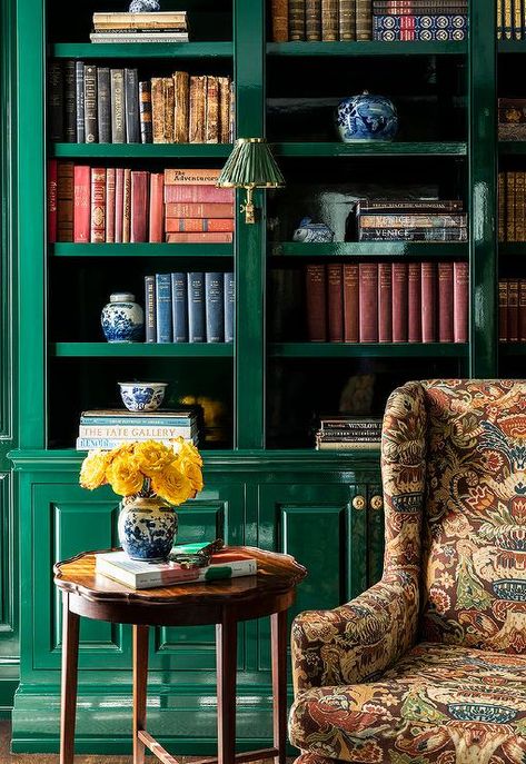 Living room features a glossy emerald green built-in bookcase with cabinets that boast brass knobs, a brown flocked wingback chair and a round wooden accent table. Green Built In Bookcase, Bookshelf Color Ideas Paint, Timeless Homes, Calke Green, Apartment Refresh, Green Bookshelves, Academia Library, Green Library, Wooden Accent Table