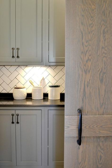 Behind a gray wash oak barn door fitted with a bronze handle, a beautiful gray laundry room features custom lit gray shaker cabinets adorning oil rubbed bronze pulls and a black quartz countertop paired with a black quartz backsplash. Grey Stained Cabinets, White Herringbone Backsplash, Gray Shaker Cabinets, Herringbone Tile Backsplash, Backsplash For White Cabinets, Backsplash With White Cabinets, Tall Kitchen Cabinets, Trendy Kitchen Backsplash, Tiles Backsplash