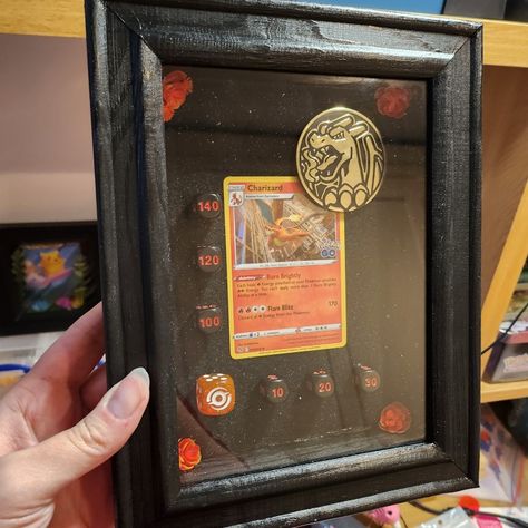 A black box frame containing a charizard pokemon card, gold charizard coin and black/orange dice. The frame's corners are accented with orange flowers. Pokemon Card Wall Art, Pokemon Cards Crafts Ideas, Trading Card Display Ideas, Pokemon Battle Scene, Pokemon Card Display, Pokemon Card Box, Nintendo Room, Pin Board Ideas, Trading Card Display