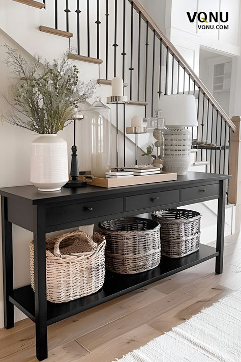 A stylish console table setup in an entryway featuring a black console table with woven baskets underneath, elegant decor items like vases and candles, and a modern staircase in the background. Black Entryway Table, Console Table Entryway, Console Entryway, Beautiful Entryways, Console Table Styling, Black Console Table, Console Table Design, Console Table Decorating, Entryway Table Decor