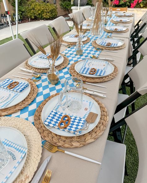 Table set up with a woven charger, gold and white cutlery, beer glass, blue diamond and white pattern table runner and napkins, meny, and beige tablecloth. Dried wheat used as centerpieces. German Party Decor, German Dinner Party, Christmas Table Setting With Blue German Plates, German Appetizers Oktoberfest Party, Woven Charger, German Beer Festival, Dried Wheat, Dinner Party Themes, Table Set Up