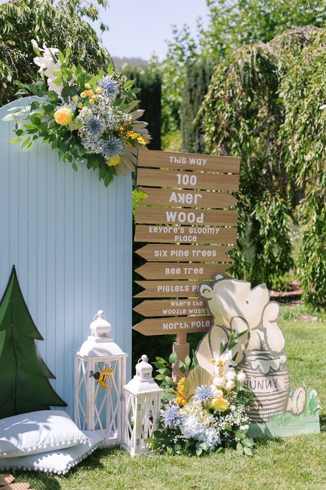 Photo backdrop setup with light blue solid wood arch, blue and yellow flowers, pastel Winnie the Pooh cardboard cutout, white lanterns, and a wooden arrow sign of the 100 Acre Woods. Diy 100 Acre Wood Sign, 100 Acre Woods Party, 100 Acre Woods Baby Shower Ideas, Hundred Acre Wood Sign, Winnie The Pooh Wedding, Wooden Arrow Sign, Brit Milah, Baby First Birthday Themes, Wooden Arrow