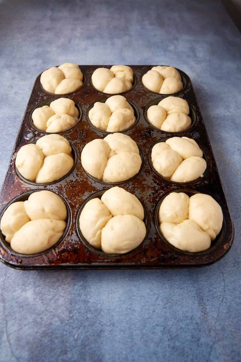 A dark metal muffin tray with twelve compartments is filled with pre-baked cloverleaf dinner roll dough divided into three segments per compartment. The tray rests on a light blue textured surface, ready to be baked. Dinner Rolls In Muffin Pan, Cloverleaf Dinner Rolls, Cloverleaf Rolls, Brioche Rolls, Dinner Roll, Homemade Breads, Homemade Rolls, Homemade Muffins, Dinner Rolls Recipe