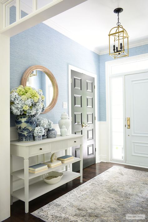 A bright and airy entryway with blue grasscloth is decorated for summer with gorgeous blue and white jars and vases arranged with gorgeous light blue and green hydrangeas. Light Blue Foyer, Hydrangea Blue Paint, Airy Entryway, Coastal Foyer, Blue Grasscloth, Blue And White Ginger Jars, Wallpapered Entryway, Spring Kitchen Decor, Room Bookshelf