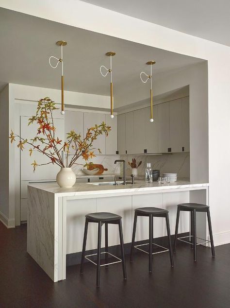 Kitchen features backless black stools at a honed marble waterfall edge peninsula illuminated by white and gold wand lights and light gray cabinets with black pulls. Kitchen Peninsula Design, White Kitchen Peninsula, Floating Sink Vanity, Peninsula Kitchen Design, Flat Front Cabinets, Black Pulls, Light Grey Kitchen Cabinets, Round Wooden Dining Table, Blue Laundry Rooms