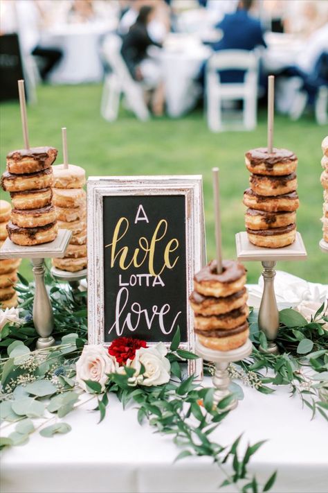 A wedding tables cape with greenery features a chalkboard sign with "A whole lotta love" written on it. Also on the table are several pedestals with multiple donuts on each pedestal, ready for guests to enjoy. Wedding Donut Dessert Table, Donut Dessert Table, Bridal Brunch Decorations, Donut Bar Wedding, Outdoor Wedding Backdrops, Donut Display, Cider Bar, Wedding Donuts, Donut Dessert