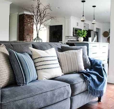 This spacious open-concept living room features a deep blue sofa decorated with soft blue and white throw pillows. The simple white walls in the living room as well as the open kitchen give a seamless look to the space. The rustic wood flooring with the brick patterns between the two rooms bring a rich and earthy component. Gray Sofa Blue Pillows, Navy Couch Neutral Pillows, Gray Couch Blue Accents, Grey Couch With Navy Blue Pillows, Slate Blue Sofa Living Room Ideas, Slate Blue Sectional, Blue Sofa Farmhouse Living Room, Accent Pillows For Blue Couch, Blue Linen Sofa Living Room