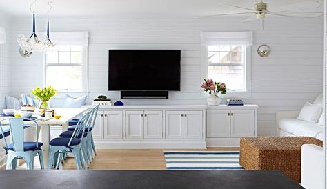 Open plan blue and white beach bungalow living room boasts a shiplap wall holding a flat panel TV mounted between two windows dressed in white roman shades and over white built in cabinets adoring vintage inset hardware lit by Pelham Moon Lights. Beach Bungalow Living Room, Built In Tv Cabinet, Bungalow Living Room, Coastal Interior, White Shiplap Wall, Wrightsville Beach, Tv Wall Decor, Cottage Living Rooms, White Shiplap