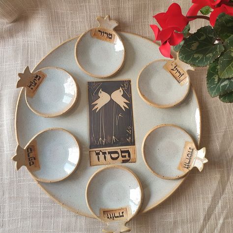 "A Passover plate with 6 matching little Pomegreantes bowls in a White glaze.  The center plate has a hand painted drawing in bronze glaze with a couple of birds, symbol of love and happiness of living.  The typography is made according to a traditional biblical Hebrew writing.  The elegant plate matches perfectly on the Passover table, a colorful addition to celebrate the holiday.  All of my works are glazed (lead-free) and fired at high temperatures (1200oC).  The items are oven, microwave, and dishwasher safe. Size: Plate diameter 11\" (28CM). Each bowl diameter 3\" (7.5CM). Embrace the slight differences in size and shape as they are a testament to the authenticity of each piece. All items are handmade in my studio, including my personal signature. Thank you for choosing my ceramic art Passover Plate, Hebrew Writing, Passover Seder Plate, Passover Table, Passover Decorations, Elegant Plates, Flower Tray, Biblical Hebrew, Shabbat Candles