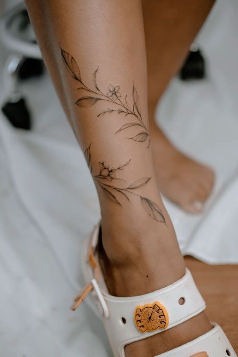 Ankle leaves tattoo bracelet leg Floral ankle tattoo on woman’s leg, with delicate leaves and blooms, next to a white sandal with an orange charm. | Sky Rye Design Leg Tattoos Women Dainty, Women Tattoos Ankle, Western Ankle Tattoos For Women, Fine Line Floral Wrap Around Tattoo, Vine Tattoo Leg Sleeve, Flower Wrap Around Tattoo Leg, Earthy Leg Tattoo, Ankle Wrap Tattoo For Women, Ankle Tattoo Wrap Around