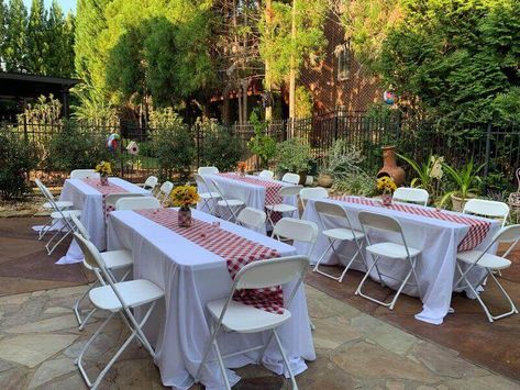 Set up charming outdoor party tablescapes with lovely decorations. Adorn the tables with white tablecloths and spread red and white checkered runners on top imparting a stunning allure to the decor. Make an attractive centerpiece by arranging sunflowers in glass vases embellished with red checkered ribbons for a gorgeous coordinated appeal. Red And White Outdoor Party, Red And White Checkered Tablecloth, Outdoor Party Table Decor, Outdoor Party Table, Table Runner Flowers, Event Decor Ideas, Party Decorations Table, Church Picnic, Picnic Table Covers