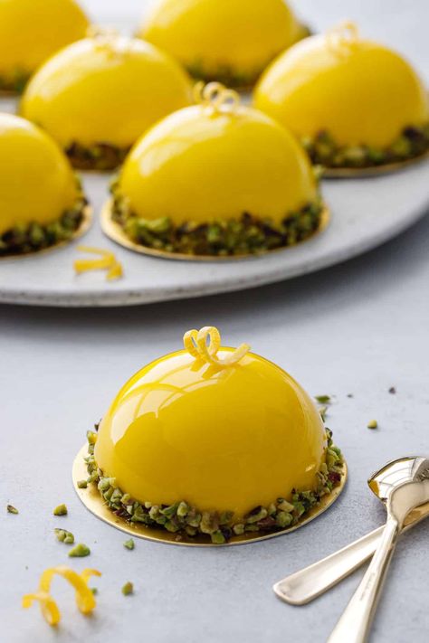 Bright yellow mirror-glazed dome shaped mousse cakes with pistachios around the edges, one cake in front and a plate of more in the background. Mini Dome Dessert, Lemon Shaped Dessert, Mousse Cake Decoration Ideas, Lemon Mousse Cake Recipe, Entremet Dessert, Lemon Mousse Cake, Meyer Lemon Curd, Mirror Glaze Recipe, Peach Mousse