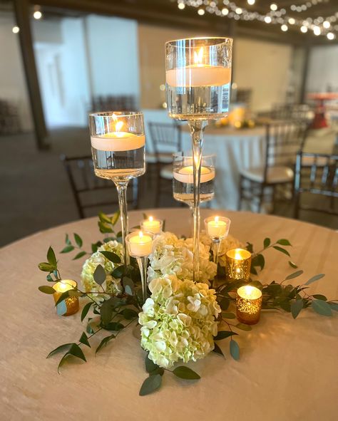 Centerpiece trends are still showing lots of candlelight and greenery. We added some hydrangeas for an added pop and are totally loving the look! The stemmed floating candles is a nice alternative to the classic cylinder look. What do you think! . . . . #trending #greenery #cnyweddings #weddingplanning #syracuseevents #nyweddings #upstatnyweddings #syracusewedding #skyarmory #syracuseweddingvenue #weddinginspiration #weddingdecor Candle Centerpiece Wedding Round Table, Candle Flower Table Decor, Floating Candles And Hydrangeas, Cylinder Vases With Floating Candles And Greenery, Candle And Hydrangea Centerpiece, Greenery Centerpiece Wedding, Floating Tea Candles, Wedding Table Decorations Elegant, Votive Centerpieces