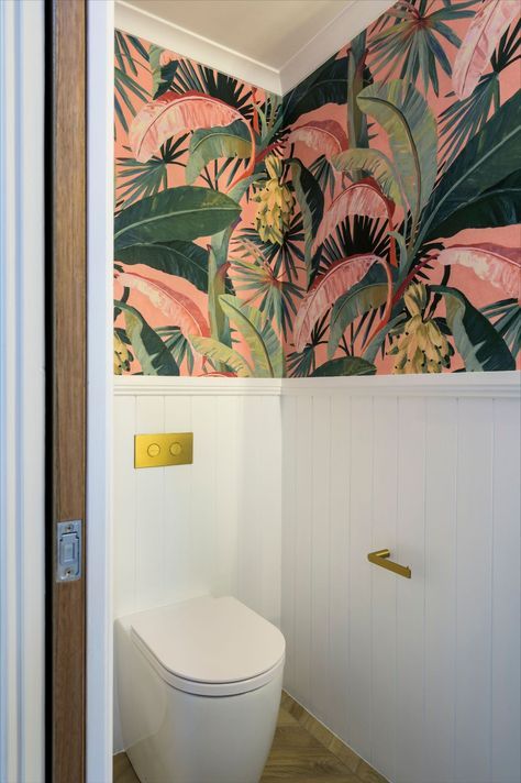 Fun Maximalism Powder Room | Maximalists and traditionalist unite in this little slice of the tropics. Herringbone laid wood-look tiles and VJ panelling speak of timeless style, while the pink and green banana palm wallpaper bursts with colour and unique expression. There’s so much to love in this separate toilet; in-wall cistern with plated buttons, brushed gold tap ware, cement Nood Co. hand basin and a stage stealing wall sconce. Bathroom Ideas Quirky, Toilet With Panelling And Wallpaper, Colourful Bathroom Wallpaper, Small Toilet With Wallpaper, Colourful Small Toilet Room, Pink And Green Downstairs Toilet, Bold Toilet Room, Tropical Wallpaper Powder Room, Colorful Toilet Design