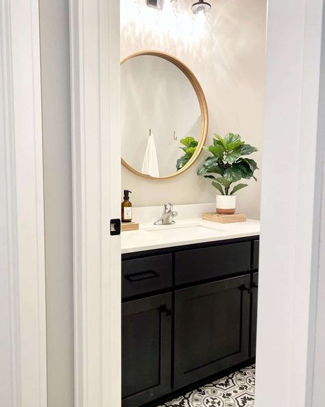 This bright bathroom uses vanity lighting to showcase the bathroom mirror. The circular mirror is complete with a wooden frame and anchored to an off-white wall. Black, white, and gray floor tile contrasts with the black vanity. Black Vanity Bathroom Floor Ideas, Black And White Flooring, Blue Kitchen Designs, Gray Floor, Grey Floor Tiles, Black Vanity Bathroom, Off White Walls, Bright Bathroom, White Tile Floor