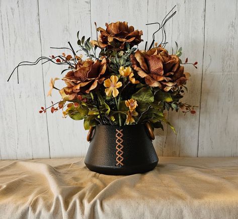 Large elegant arrangement created in a rustic black metal cauldron with suede leather lacing on the front, and leather straps with wood rings on the sides. This arrangement is unique in that it combines high end elegance with rustic and a touch of western flair. Featured are very high quality brown roses with tan highlights, gold floral accents, assorted greenery, fall bog pimpernel accents, and wood accent stems. There is no way to describe how absolutely gorgeous this arrangement is in person!  Dimensions for overall arrangement: 15" High x 15" Wide Dimensions for metal vase only: 5" High x 8" Wide Peach Fall Decor, Western Floral Centerpieces, Diy Western Flower Arrangement, Western Fake Flower Arrangements, Western Floral Arrangements Home, Longhorn Floral Arrangement, Tuscan Floral Arrangements, Silk Floral Centerpiece, Rustic Flower Arrangements