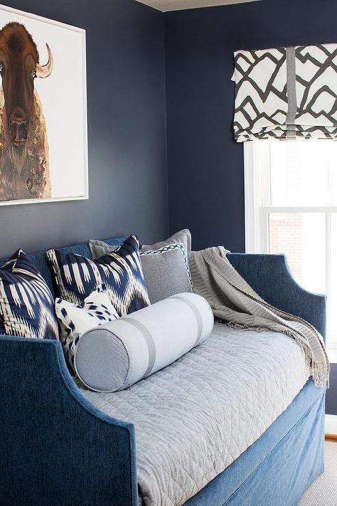 Well designed contemporary boy's bedroom features a dark blue velvet skirted daybed dressed in gray bedding and topped with a white and silver bolster pillow, blue ikat pillows, gray pillows with white borders, and a gray throw blanket positioned under an American bison canvas mounted on dark blue walls framing a window covered in a roman shade in Zimba Charcoal Fabric. Daybed Room Ideas, Grey Boys Rooms, Blue Daybed, Blue Velvet Skirt, Daybed Room, Bedroom Dark, Blue Dresser, Dark Blue Velvet, Dark Blue Walls