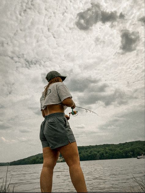 Girl fishing in white crop top and workout shorts in nature outside on a cloudy day. Cute Fishing Outfit For Women Summer, Fishing Date Outfit, Fishing Outfits For Women Summer, Cute Fishing Outfits, Fishing Outfits For Women, Lake Fits, Lake Outfits, Outdoorsy Aesthetic, Fishing Outfit