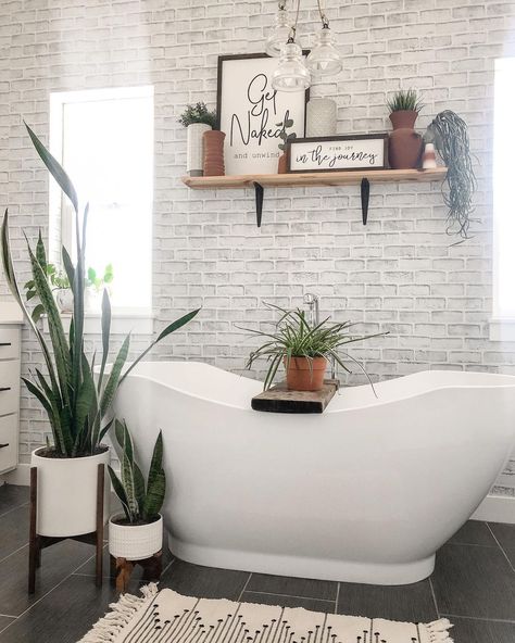 This bathroom with whitewashed brick walls features a white freestanding oval soaking tub with rustic wood bath tray. A light wood and black metal shelf is mounted above the bathtub between two white windows. Soaking Tub Decor, Small Bathroom Look Bigger, Get Naked Sign, Black Bathroom Floor, Wood Bath Tray, White Subway Tile Shower, Brick Bathroom, White Wash Brick, White Tub