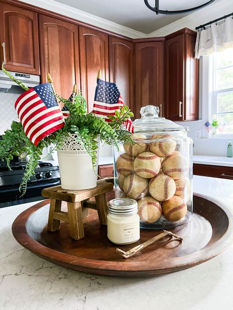 Wood bowl on a kitchen island with large glass container filled with old baseballs. Small container on a a wood stool riser sits next to the baseballs with mini American flags and faux ferns.