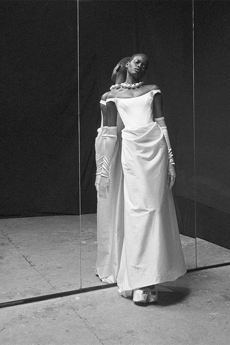 Black and white photo of a model wearing the Vivienne Westwood Grand Camille bridal gown. The gown features an off-the-shoulder neckline, structured bodice, and draped detailing. The model poses elegantly in front of a mirror, wearing long satin gloves and a statement pearl necklace, creating a dramatic and timeless bridal look. The Originals Camille, Vivienne Westwood Bridal, Vivienne Westwood Wedding Dress, Vivienne Westwood Wedding, Sustainable Wedding Dress, Camille Dress, Wedding Dress Brands, The Vivienne, Taffeta Dress