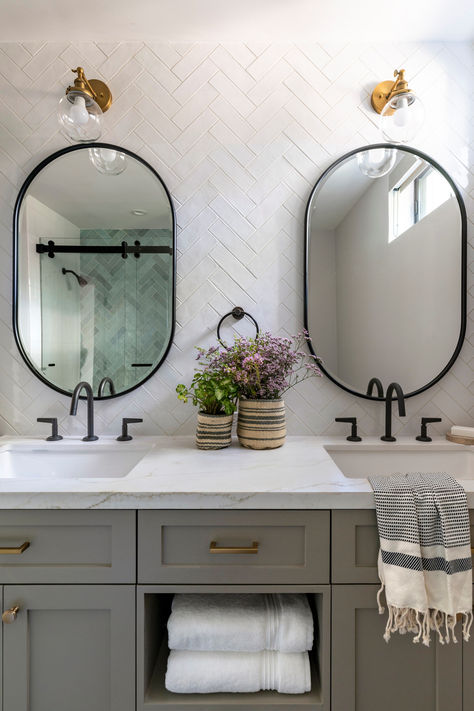 We are daydreaming of this perfect bathroom renovation. The muted gray color of the custom cabinetry with white countertops creates a lovely contrast with the black plumbing fixtures. Adding to the design, we incorporated two mirrors with an organic shape and a black edge detail, harmonizing with the plumbing. To complete the look, we decided to place contrasting brass sconces for both mirrors. We believe the result of this bathroom is truly charming! Powder Room Ideas Grey Vanity, Brass And Black Bathroom Hardware, Farmhouse Bathroom With Black Fixtures, Farmhouse Plumbing Fixtures, Coastal Bathroom Black Fixtures, Bathroom Vanity With Grey Tile, Master Bath Gold Hardware, Black Bathroom With Gold Fixtures, Bathroom With Black Fixtures And Wood