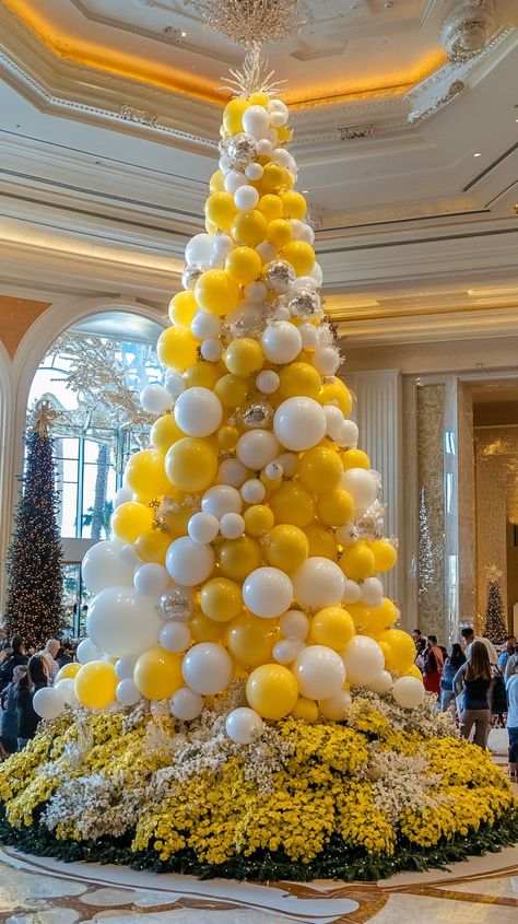 A grand Christmas tree adorned with yellow and white ornaments, surrounded by flowers and a balloon art installation in a Yellow Christmas Aesthetic, Gold Christmas Tree Ideas, Poinsettia Tree, Floral Christmas Tree, Las Vegas Hotel, Rosé Christmas, Christmas 2025, Vibrant Bouquet, Christmas Planters