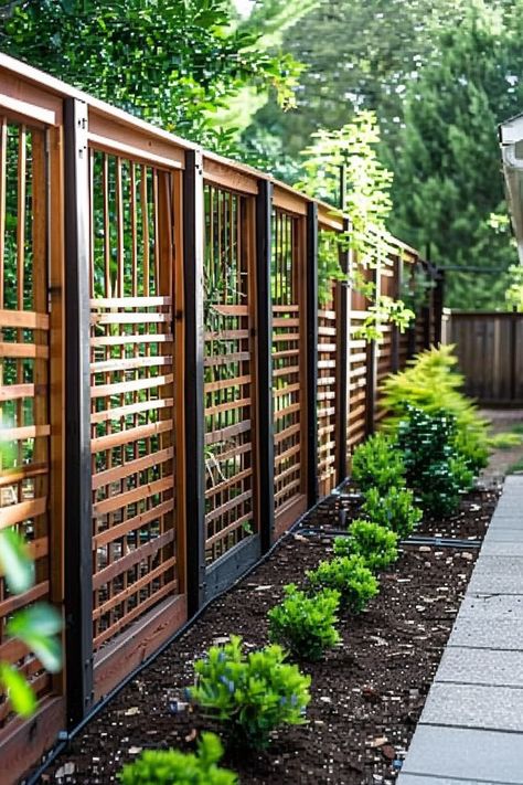 Wooden fence with metal posts lines a garden with small green plants, next to a paved path, against a backdrop of trees.