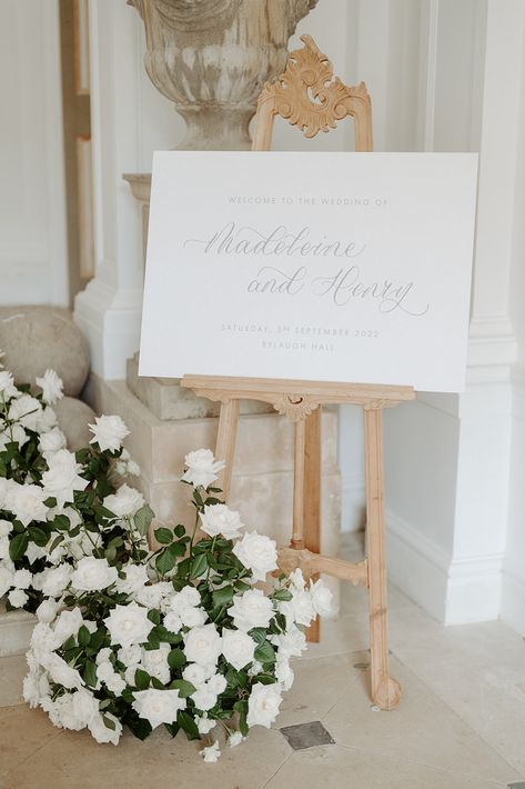 A landscape wedding welcome sign printed on a white background with grey calligraphy and text. The sign is displayed on an ornate wooden carved easel at the bottom of a small flight of stairs. An arrangement of white roses is displayed around the foot of the easel and a large stone urn stands behind the easel. Wedding Signage Table, Wedding Table Signage, Sign Easel, Suzanne Neville, Event Signage, Wooden Easel, Event Sign, Wedding Hire, Easels