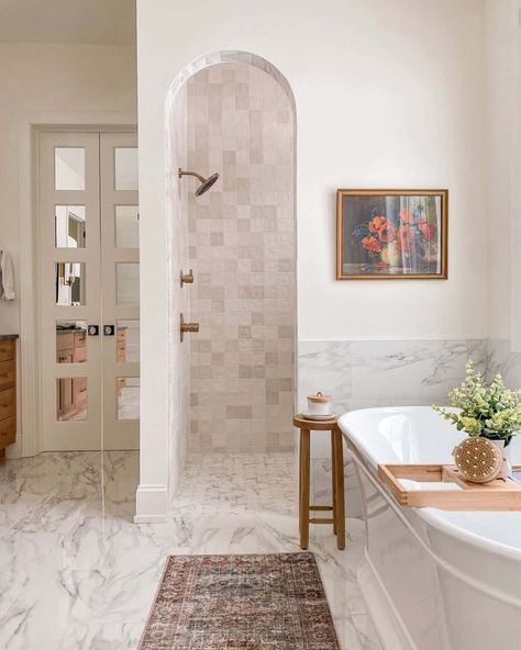 This cream and white bathroom features a hidden doorless walk-in shower behind a wall partition, a white tub, and marble flooring. A small table accessorized bath salts rest near the tub. A dark, traditional runner stretches along the floor below the soaking tub. Hidden Shower, Small Master Bath, Doorless Shower, Master Bath Shower, Window In Shower, White Tub, Master Shower, Cottage Bathroom, Master Bath Remodel