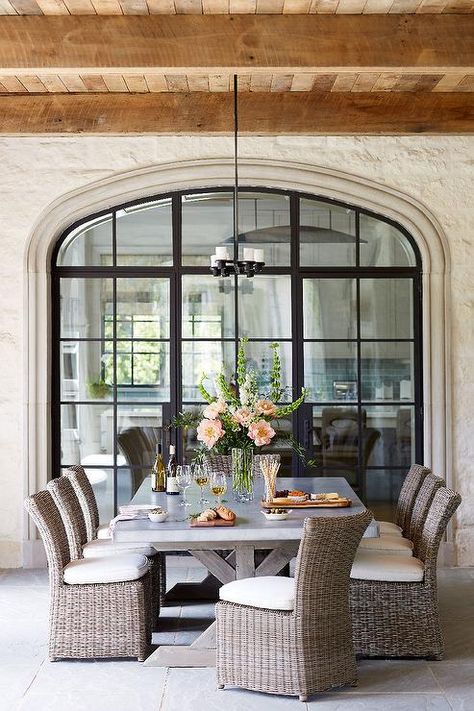 In front of iron framed patio doors, a linear iron chandelier hangs from a covered patio plank ceiling above a Restoration Hardware Salvaged Wood & Weathered Concrete X-Base Rectangular Dining Table surrounded by wicker slipper chairs placed on concrete pavers. Spanish Style Kitchen Backsplash, Modern Spanish Style Kitchen, Spanish Kitchen Design, Modern Spanish Style, Spanish Style Kitchen, Bistro Furniture, Mediterranean Home Decor, Arched Doors, Spanish Style Homes