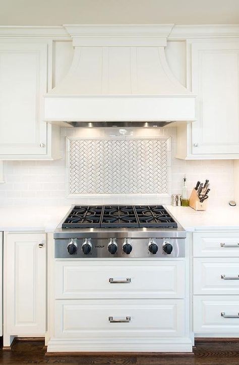 A white paneled French kitchen hood stands over a white herringbone cooktop backsplash and an integrated Wolf cooktop situated above stacked pot and pan drawers. Cooktop Backsplash, Wolf Cooktop, Kitchen Hood Ideas, Kitchen Vent Hood, Kitchen Vent, French Kitchen Decor, Kitchen Hood, Kitchen Backsplash Designs, White Herringbone