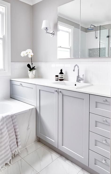 Beautiful gray and white bathroom is fitted with a gray washstand adorning polished nickel knobs and a white quartz countertop holding an overmount sink with a polished nickel gooseneck faucet in front of white subway backsplash tiles tiled with white pencil tiles fixed beneath a vanity mirror flanked by nickel sconces mounted on a gray wall. Grey Bathroom Paint, Grey Bathroom Cabinets, Makeover Kamar Mandi, Ideas Baños, Gray And White Bathroom, Grey Bathroom Tiles, Grey Bathroom Vanity, Sconces Bathroom, Bad Inspiration