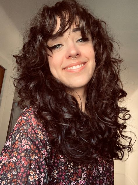 Girl smiling in selfie photo. Long brown curly hair with bangs, blue eyes with black winged eye-liner, septum ring in nose. Wearing a dark floral top. Med Length Curly Hair With Bangs, Butterfly Haircut With Bangs Curly Hair, Loose Curly Hair With Bangs, Layered Chunky Curls With Bangs, Shoulder Length 2b Hair, Curly Layers With Bangs, Medium Curly Shag With Bangs, Curly Shag Haircut With Bangs, Curly Hair Cuts Bangs