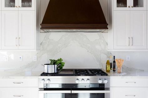 A brown vent hood is mounted above a stainless steel oven range to a bookmatched marble backsplash between white cabinets stacked under glass display shelves. Miele Range, White Kitchen Paint Colors, Oven Range Hood, White Kitchen Paint, Glass Display Shelves, Kitchen Vent Hood, Oven Hood, White Marble Kitchen, Kitchen Vent