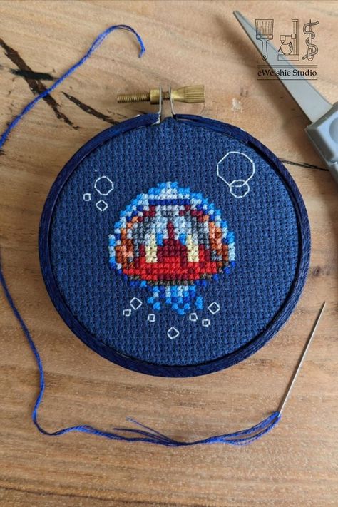 A finished cross stitch of a small jellyfish with red insides on blue cross fabric. Jellyfish Cross Stitch, Small Jellyfish, Floating In The Ocean, Embroidery Floss Crafts, Cute Jellyfish, Copper Light, Blue Jellyfish, Graph Paper Art, Bead Sprite