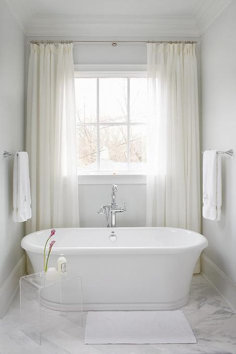 White sheer curtains dress a window placed above a freestanding oval bathtub paired with a polished nickel floor-mount tub filler alongside a small lucite waterfall accent table situated on a white marble herringbone pattern tiled floor. Small Freestanding Tub, Bathroom Window Treatments, Simple Bathroom Remodel, Small Window Curtains, White Sheer Curtains, Bathroom Tub, Diy Bathroom Remodel, Bathroom Windows, Trendy Bathroom